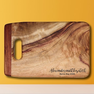 以下が含まれることがあります： ハンドル付きの長方形の木製のまな板。まな板は自然な木目模様と濃い茶色の縁取りがあります。まな板には「Alexander and Elizabeth Byron Bay 2026」という文字が刻まれています。