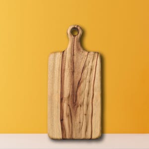 May include: A rectangular wooden cutting board with a handle and a hole for hanging. The board is made of light brown wood with darker brown grain patterns. The handle is rounded and has a hole for hanging. The background is a solid yellow color.