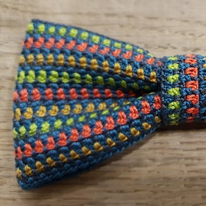 Puede incluir: Una pajarita colorida con un patrón azul, verde, naranja y dorado. La pajarita está hecha de un tejido tejido y tiene una superficie texturizada.