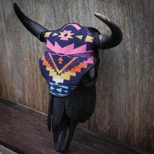 May include: A black bull skull with large, curved horns. The skull is adorned with a colorful fabric mask featuring geometric patterns in pink, blue, orange, and yellow. The skull is mounted on a wooden surface, creating a rustic, decorative piece.