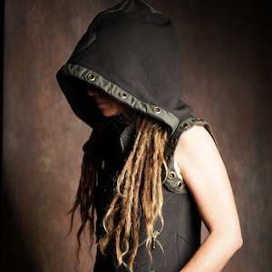 May include: A black hooded vest with a zipper closure and multiple grommets. The vest has a green trim and is worn over a black shirt. The person wearing the vest has long, blonde dreadlocks.