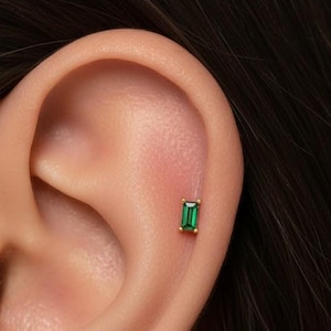 May include: A close-up of an ear with a small, rectangular emerald-green stud earring set in gold. The earring is positioned on the upper ear cartilage. The skin tone is light, and the hair is dark.