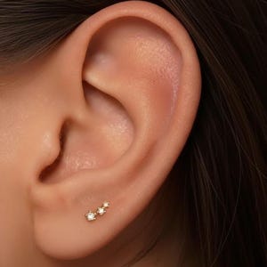 May include: A close-up of an ear with a small, gold earring. The earring features three small, sparkling gemstones arranged in a line. The skin around the ear is smooth and has a warm, neutral tone.