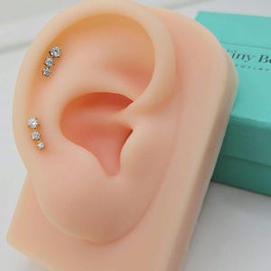 May include: A display of ear piercings featuring three small, sparkling, clear-stone earrings. The earrings are set in gold-colored metal and are arranged in a cascading design. The ear is displayed on a flesh-toned mannequin.
