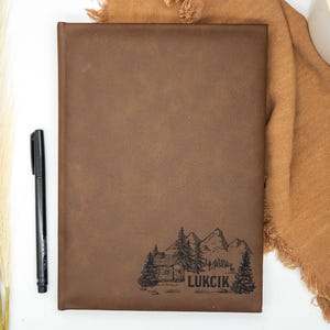May include: A brown leather journal with the word "LUKCIK" and a mountain and cabin illustration. A black pen rests beside the journal. The journal is on a white surface with decorative elements.