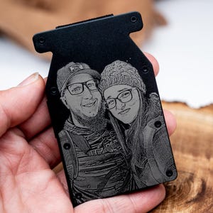 Puede incluir: Una cartera de metal negro con un retrato grabado con láser de una pareja que lleva gafas y sombreros. La cartera se sostiene en una mano y tiene forma rectangular con esquinas redondeadas.