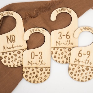 Puede incluir: Cuatro divisores de ropa de bebé de madera con la parte superior curvada. Los divisores están etiquetados como "Recién nacido", "0-3 meses", "3-6 meses" y "6-9 meses". Cada uno tiene un patrón floral en la parte inferior. Los divisores están sobre una tela marrón.