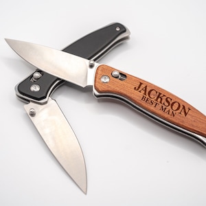 May include: A pocket knife with a wooden handle engraved with the text "JACKSON BEST MAN". The knife has a black handle and a silver blade.
