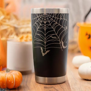 May include: Black stainless steel tumbler with a spiderweb and bats design. The tumbler is surrounded by Halloween decorations, including pumpkins and candy.
