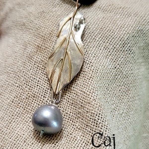 May include: Silver-tone leaf pendant necklace with gold-tone accents and a gray pearl drop. The leaf design features intricate veining and two small, clear stones. The necklace hangs from a black cord.
