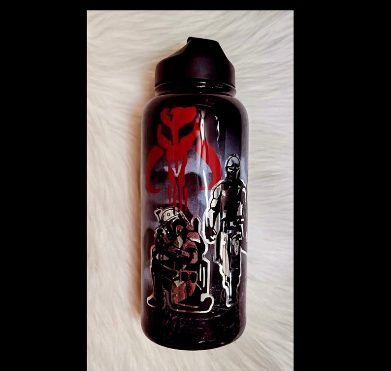 Peut inclure: Gourde noire avec un bouchon noir. Elle pr&eacute;sente un graphisme rouge et blanc d'un cr&acirc;ne Mandalorien et de deux personnages. La gourde a un fond sombre et enfum&eacute; et un dessus noir.