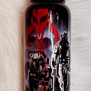 Peut inclure: Gourde noire avec un couvercle noir, orn&eacute;e d'images rouges et argent&eacute;es de personnages et de symboles d'une s&eacute;rie de science-fiction populaire. Le design comprend un embl&egrave;me rouge et des personnages en armure.