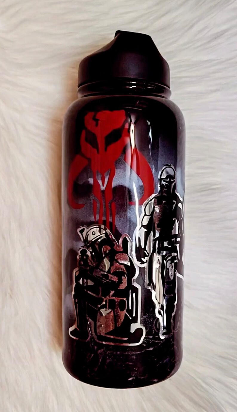 Peut inclure: Gourde noire avec un couvercle noir, orn&eacute;e d'images rouges et argent&eacute;es de personnages et de symboles d'une s&eacute;rie de science-fiction populaire. Le design comprend un embl&egrave;me rouge et des personnages en armure.