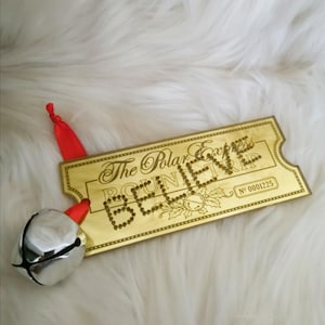 May include: A gold-colored ticket-shaped ornament with the words "BELIEVE" spelled out in small dots. The text "The Polar Express" is above the word "BELIEVE". A silver bell with a red ribbon is attached to the ornament.