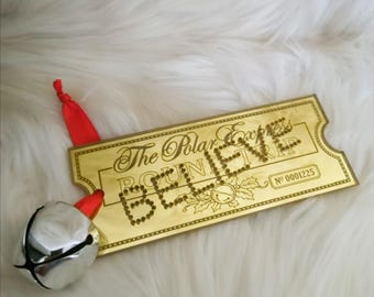 May include: A gold-colored ticket-shaped ornament with the words "BELIEVE" spelled out in small dots. The text "The Polar Express" is above the word "BELIEVE". A silver bell with a red ribbon is attached to the ornament.