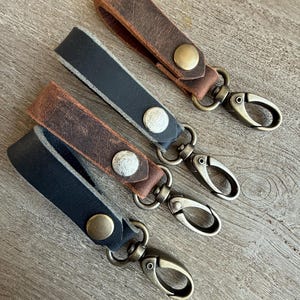 May include: Three leather keychains with antique brass swivel clips and snap closures. The keychains are brown, black, and brown with a lighter brown trim.