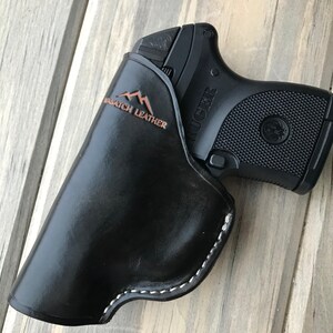 Peut inclure: Un &eacute;tui en cuir noir avec des d&eacute;tails de couture blanche. L'&eacute;tui est con&ccedil;u pour contenir un petit pistolet noir avec le mot "RUGER" imprim&eacute; sur le c&ocirc;t&eacute;. L'&eacute;tui porte l'inscription "Mountain Stitch Leather" sur le c&ocirc;t&eacute;.