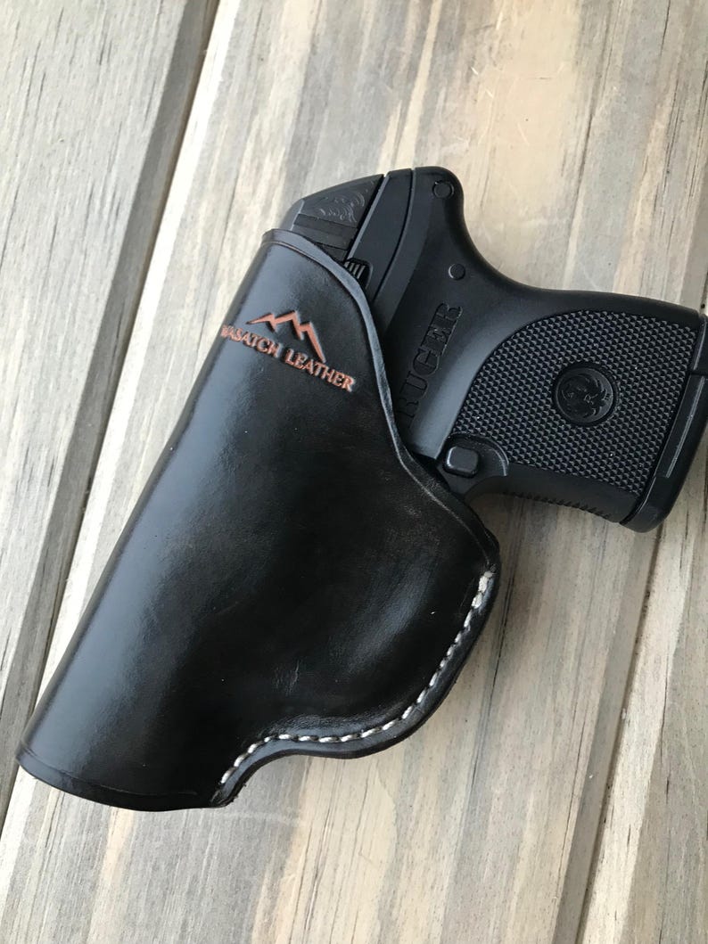 Peut inclure: Un &eacute;tui en cuir noir avec des d&eacute;tails de couture blanche. L'&eacute;tui est con&ccedil;u pour contenir un petit pistolet noir avec le mot "RUGER" imprim&eacute; sur le c&ocirc;t&eacute;. L'&eacute;tui porte l'inscription "Mountain Stitch Leather" sur le c&ocirc;t&eacute;.