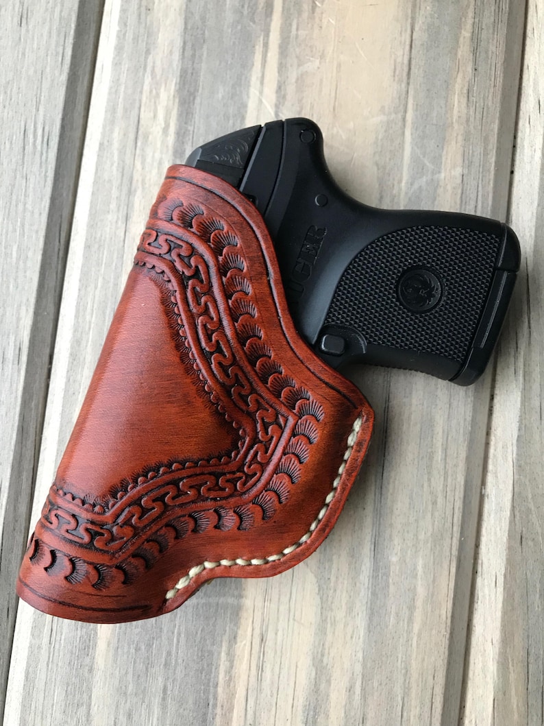 Peut inclure: Un &eacute;tui en cuir marron avec des d&eacute;tails et des coutures complexes. L'&eacute;tui est con&ccedil;u pour contenir une petite arme &agrave; feu noire.