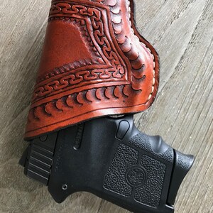 Peut inclure: Un &eacute;tui en cuir marron avec des motifs et des coutures complexes. L'&eacute;tui est con&ccedil;u pour contenir un petit pistolet noir. Le pistolet a une poign&eacute;e textur&eacute;e et une finition noire.