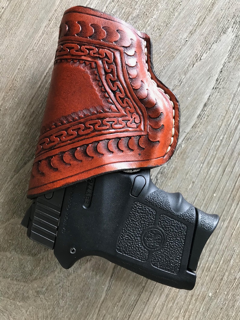 Peut inclure: Un &eacute;tui en cuir marron avec des motifs et des coutures complexes. L'&eacute;tui est con&ccedil;u pour contenir un petit pistolet noir. Le pistolet a une poign&eacute;e textur&eacute;e et une finition noire.