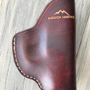 Peut inclure: Un &eacute;tui en cuir marron avec des d&eacute;tails de couture blancs et le texte "WASATCH LEATHER" et un logo de montagne.