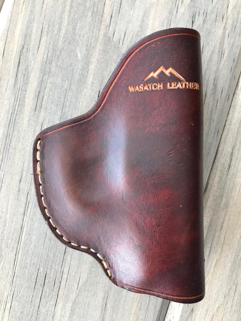 Peut inclure: Un &eacute;tui en cuir marron avec des d&eacute;tails de couture blancs et le texte "WASATCH LEATHER" et un logo de montagne.