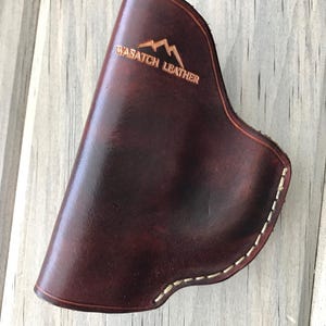 Peut inclure: Un &eacute;tui en cuir marron avec des coutures blanches et le texte "Wasatch Leather" en relief sur le devant.