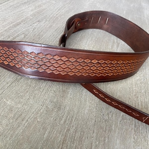 May include: A brown leather guitar strap with a diamond pattern embossed on the front. The strap is made from high-quality leather and is designed to be comfortable and durable.