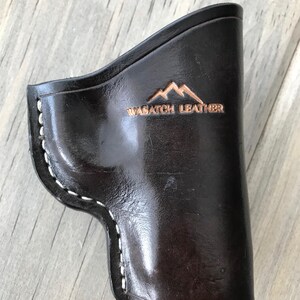 Peut inclure: Un &eacute;tui en cuir noir avec des coutures blanches et le texte "WASATCH LEATHER" en relief sur le devant. L'&eacute;tui a un logo de montagne au-dessus du texte.