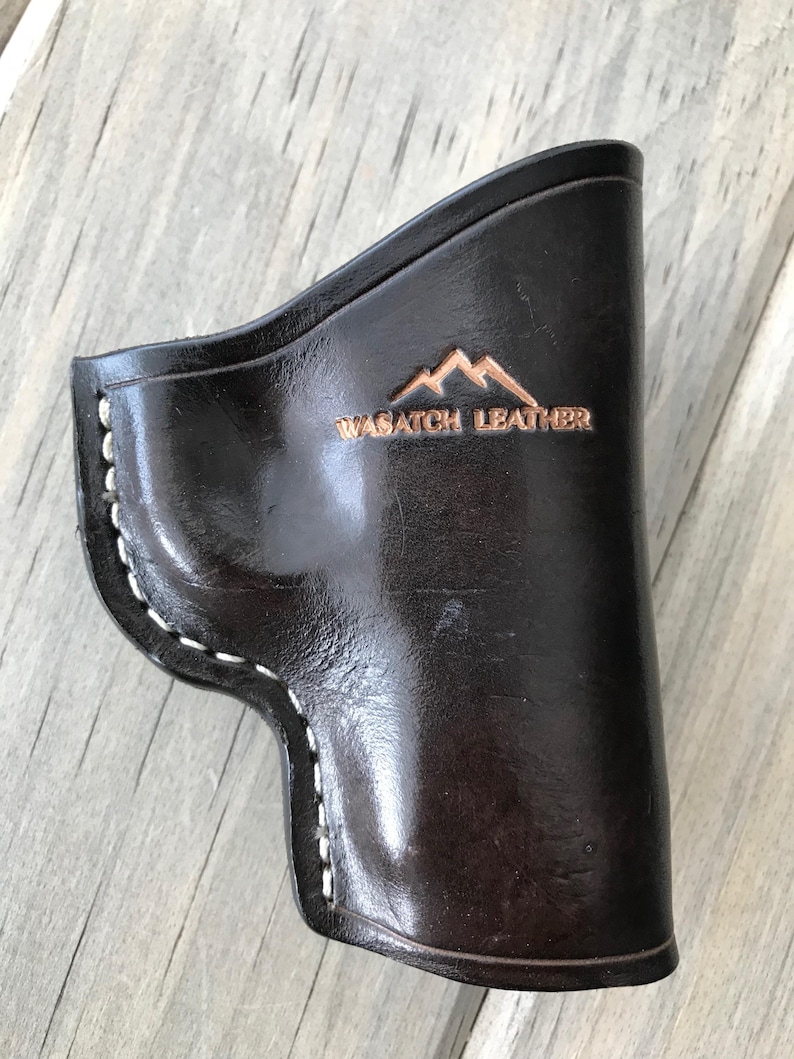 Peut inclure: Un &eacute;tui en cuir noir avec des coutures blanches et le texte "WASATCH LEATHER" en relief sur le devant. L'&eacute;tui a un logo de montagne au-dessus du texte.