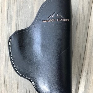 Peut inclure: Un &eacute;tui en cuir noir avec des coutures blanches et le texte "WASATCH LEATHER" en relief sur le devant.