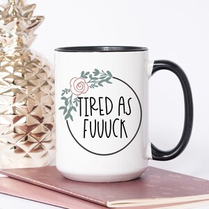 May include: White ceramic mug with a black rim and handle. The mug has a floral design with a pink rose and green leaves around the text "TIRED AS FUUVCK".