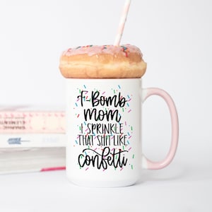 May include: White ceramic mug with a pink handle and the text "F-Bomb Mom I Sprinkle That Shit Like Confetti" in black lettering with colorful confetti sprinkles around the text. A pink frosted donut with colorful sprinkles sits on top of the mug.