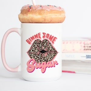 Peut inclure: Une tasse en céramique blanche avec une anse rose. La tasse est décorée d'une bouche de lèvres en imprimé léopard rose et noir avec le texte "Gimme Some Sugar" en rose.