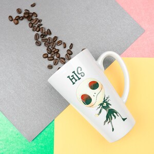 Może przedstawiać: Biały ceramiczny kubek do latte z kresk&oacute;wkowym wzorem postaci Jacka Skellingtona z filmu Koszmar przed świętami. Kubek ma napis "HIS" nadrukowany na boku. Wok&oacute;ł kubka rozrzucone są ziarna kawy.