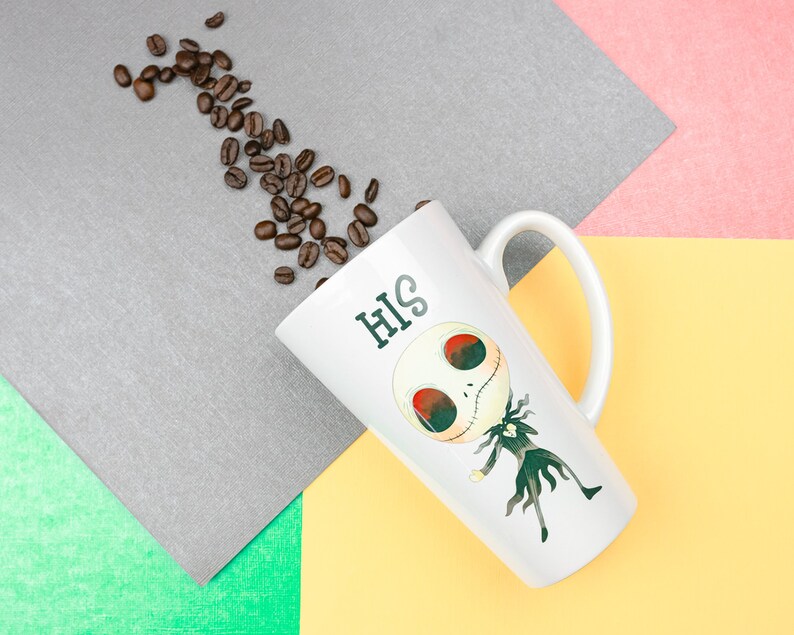 Może przedstawiać: Biały ceramiczny kubek do latte z kresk&oacute;wkowym wzorem postaci Jacka Skellingtona z filmu Koszmar przed świętami. Kubek ma napis "HIS" nadrukowany na boku. Wok&oacute;ł kubka rozrzucone są ziarna kawy.