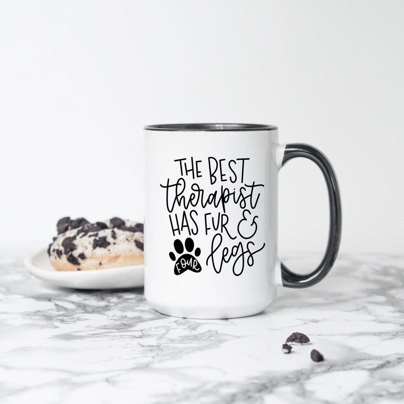 Puede incluir: Taza de cer&aacute;mica blanca con borde y asa negros. La taza tiene una huella de pata negra con la palabra "cuatro" dentro y el texto "El mejor terapeuta tiene pelaje y patas".