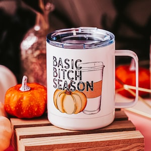 Peut inclure: Une tasse en céramique blanche avec un motif noir et blanc d'une citrouille et d'un gobelet à café. Le texte sur la tasse est "Basic Bitch Season".