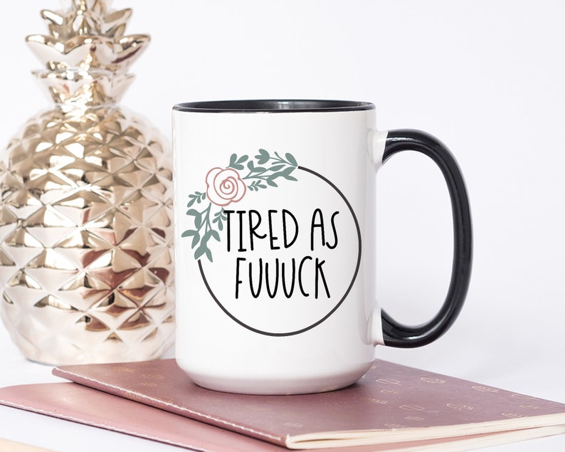 May include: White ceramic mug with a black rim and handle. The mug has a floral design with a pink rose and green leaves around the text "TIRED AS FUUVCK".