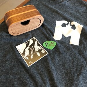 Könnte beinhalten: Ein dunkelgraues T-Shirt mit einer weißen Grafik eines stilisierten Buchstabens M. Eine Holzkiste mit Deckel und ein schwarz-weißer Aufkleber befinden sich auf dem T-Shirt. Ein grüner Gitarrenplektrum mit den Buchstaben JM befindet sich auf dem T-Shirt.