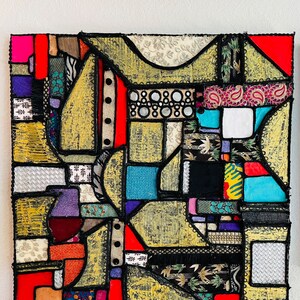 Op de afbeelding: Abstract kunstwerk gemaakt van kleurrijke stoffen die in een geometrisch patroon aan elkaar zijn genaaid. De stof is een mix van texturen en patronen, waaronder paisley, bloemen en geometrische prints. Het stuk is omlijst met zwarte kant en heeft een gouden achtergrond.