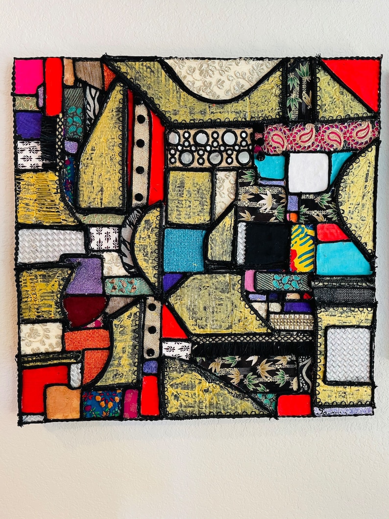 Op de afbeelding: Abstract kunstwerk gemaakt van kleurrijke stoffen die in een geometrisch patroon aan elkaar zijn genaaid. De stof is een mix van texturen en patronen, waaronder paisley, bloemen en geometrische prints. Het stuk is omlijst met zwarte kant en heeft een gouden achtergrond.