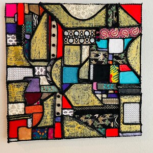Op de afbeelding: Abstract kunstwerk gemaakt van kleurrijke stoffen die in een geometrisch patroon aan elkaar zijn genaaid. De stof is een mix van texturen en patronen, waaronder paisley, bloemen en geometrische prints. Het stuk is omlijst met zwart en heeft een gouden achtergrond.