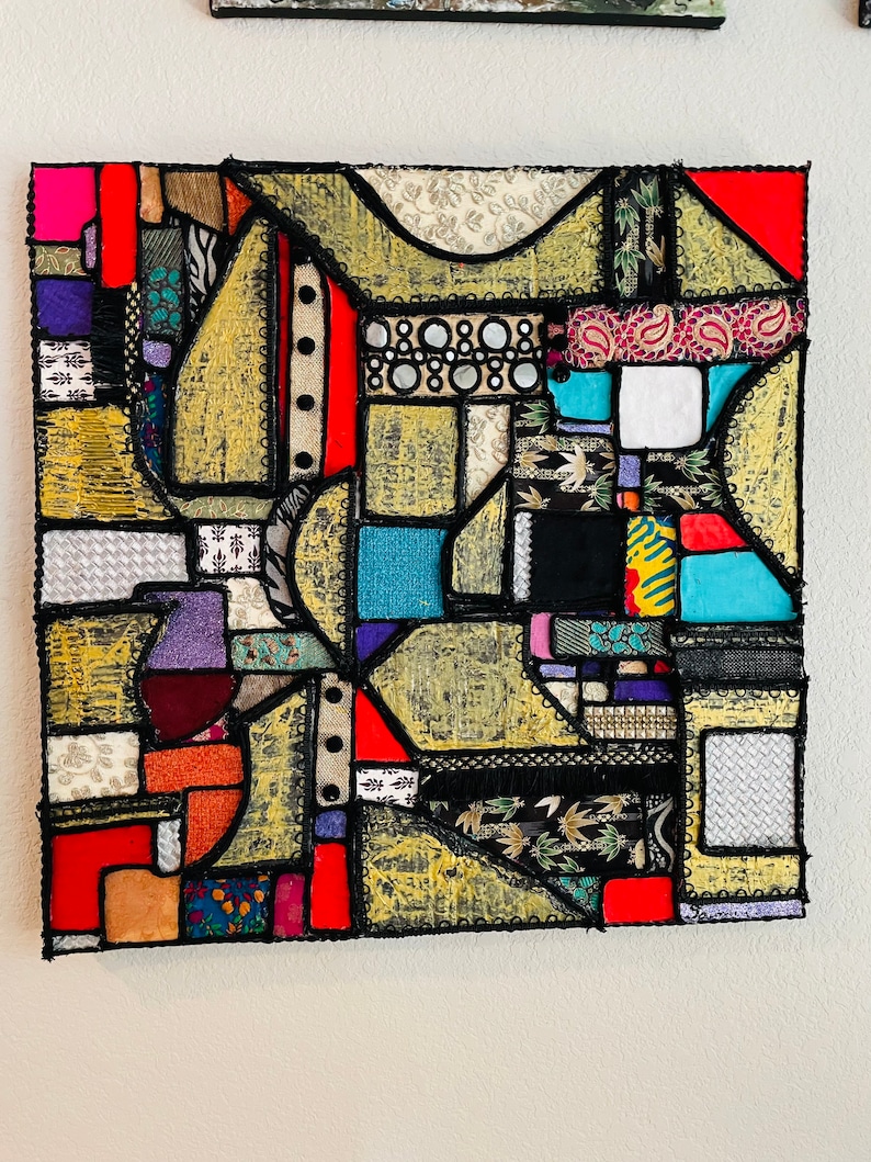 Op de afbeelding: Abstract kunstwerk gemaakt van kleurrijke stoffen die in een geometrisch patroon aan elkaar zijn genaaid. De stof is een mix van texturen en patronen, waaronder paisley, bloemen en geometrische prints. Het stuk is omlijst met zwart en heeft een gouden achtergrond.