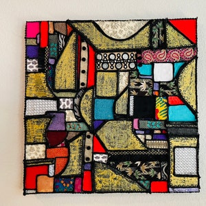 Op de afbeelding: Een kleurrijk abstract kunstwerk gemaakt van stoffen resten die in een geometrisch patroon aan elkaar zijn genaaid. Het werk heeft een verscheidenheid aan kleuren, waaronder rood, blauw, groen, geel en zwart. De stof is in verschillende vormen en maten geknipt, en de naden zijn zichtbaar. Het werk is ingelijst in een zwart frame.