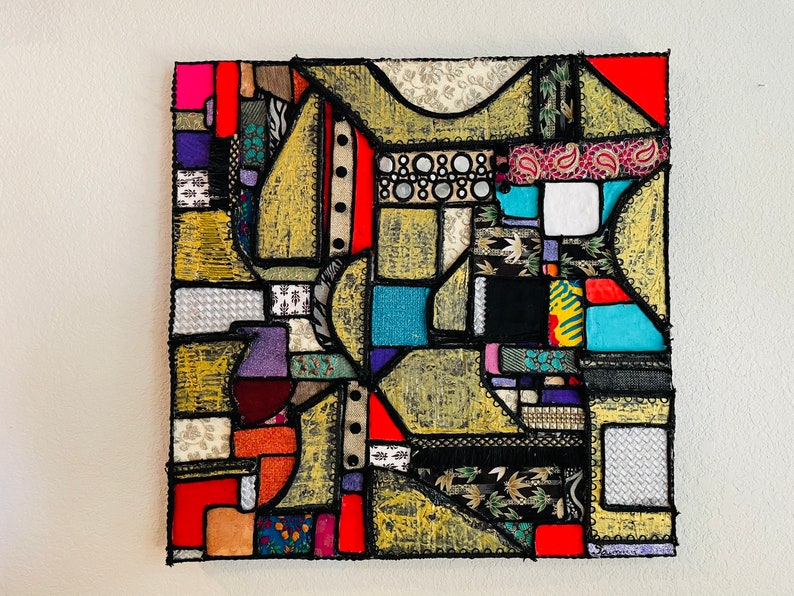 Op de afbeelding: Een kleurrijk abstract kunstwerk gemaakt van stoffen resten die in een geometrisch patroon aan elkaar zijn genaaid. Het werk heeft een verscheidenheid aan kleuren, waaronder rood, blauw, groen, geel en zwart. De stof is in verschillende vormen en maten geknipt, en de naden zijn zichtbaar. Het werk is ingelijst in een zwart frame.