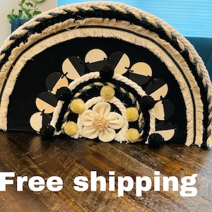 May include: A black and white macrame wall hanging with a flower design in the center. The hanging is made of rope and has a fringe border.