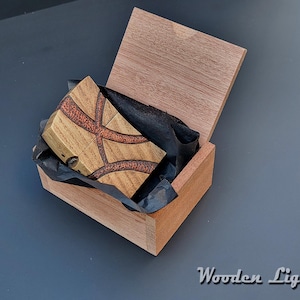 May include: A wooden lighter with a geometric design in a wooden box. The lighter features a light brown wood with a darker, reddish-brown inlay. The box is open, revealing the lighter nestled on black tissue paper. The text "Wooden Lighter" is visible.