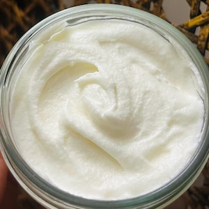 May include: A close-up shot of a clear glass jar filled with a thick, white, creamy substance. The cream has a swirled texture, suggesting a whipped or emulsified consistency. The jar is set against a blurred background.